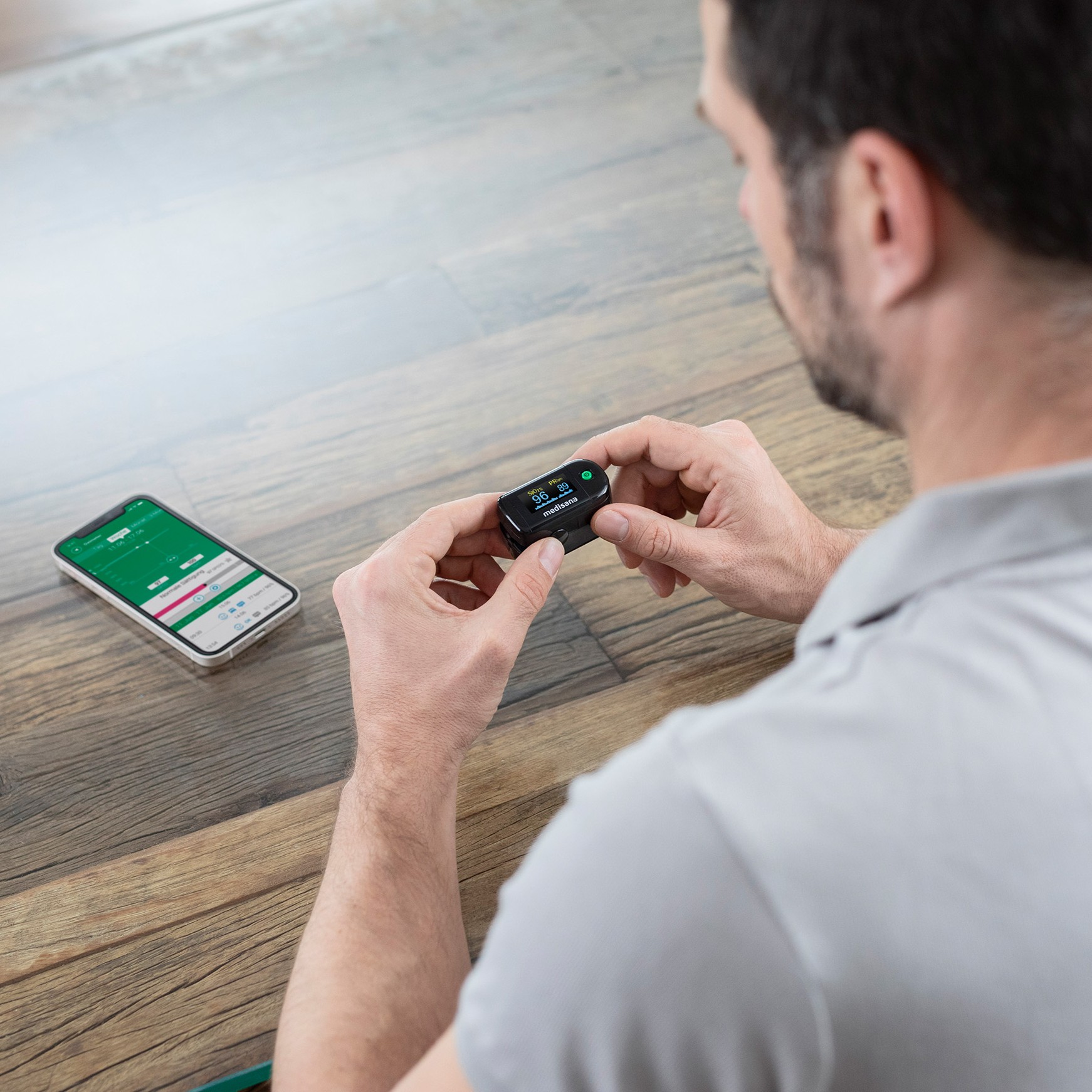A man holding a black Medisana pulse oximeter while a smartphone with the connected health app lies next to him.