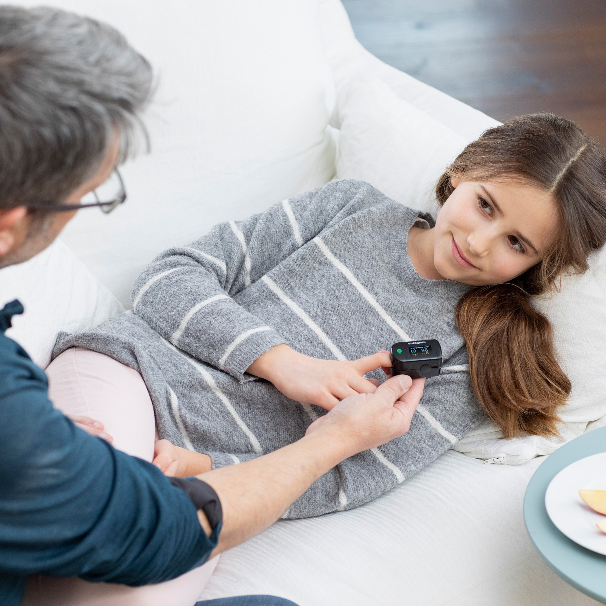 A girl lying on a sofa measuring her oxygen saturation with a black Medisana pulse oximeter, assisted by an adult.